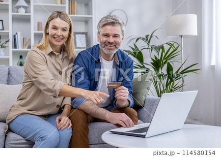 Joyful couple using a laptop and credit card for online shopping. They are sitting comfortably on a sofa in a cozy living room. Joyful couple using a laptop and credit card for online shopping. They are sitting comfortably on a sofa in a cozy living room. 114580154