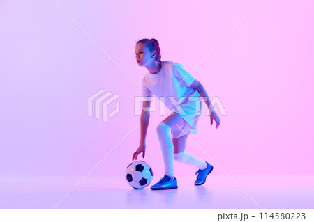 Teen girl in white uniform, football player with ball practicing against pink studio background in neon light 114580223