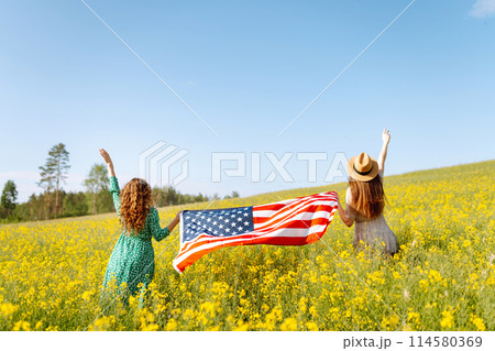 Two Young woman with american flag on blooming meadow. 4th of July. USA flag fluttering in the wind. Two Young woman with american flag on blooming meadow. 4th of July. USA flag fluttering in the wind. 114580369