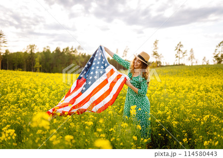 Young woman with american flag on blooming meadow. Independence Day. USA flag fluttering in the wind Young woman with american flag on blooming meadow. Independence Day. USA flag fluttering in the wind 114580443