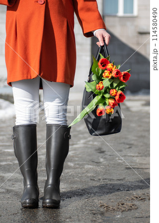 A beautiful young woman in a orange coat and rubber boots walking down the street with a bouquet of tulips A beautiful young woman in a orange coat and rubber boots walking down the street with a bouquet of tulips 114580490