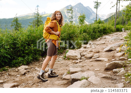 Young female traveler with hiking poles, bright backpack enjoys mountain scenery. Active lifestyle. 114583371