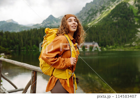 Female traveler with yellow hiking backpack against the backdrop of mountain lake with hiking poles. 114583407