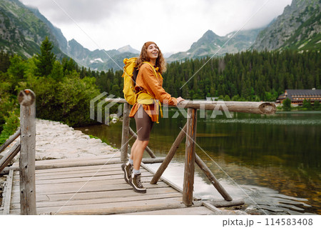 Female traveler with yellow hiking backpack against the backdrop of mountain lake with hiking poles. Female traveler with yellow hiking backpack against the backdrop of mountain lake with hiking poles. 114583408