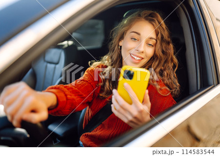 Portrait of a young woman sitting in a car in the driver's seat looking into a smartphone. 114583544