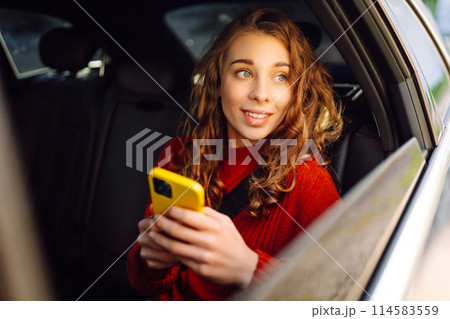 Positive young woman in casual clothes sits in the back seat of a car with a mobile phone. Positive young woman in casual clothes sits in the back seat of a car with a mobile phone. 114583559