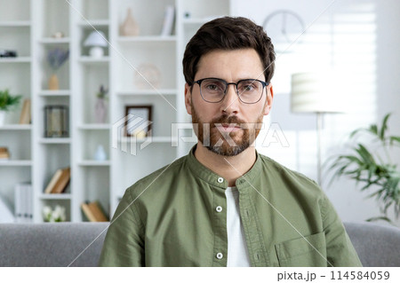 A serious man with glasses sitting in a modern home office, looking thoughtfully at the camera. Shelves with decor in the background. 114584059