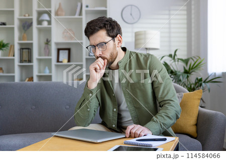 Thoughtful young man working on his laptop at home. He is thinking and planning while sitting on the sofa. 114584066