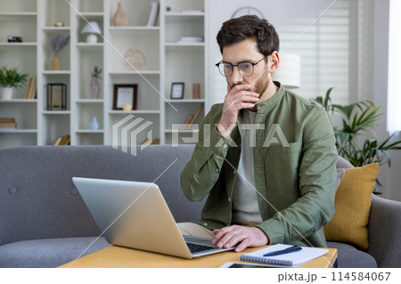 Man working from home on a laptop, thinking and analyzing projects in a modern living room setting. Man working from home on a laptop, thinking and analyzing projects in a modern living room setting. 114584067