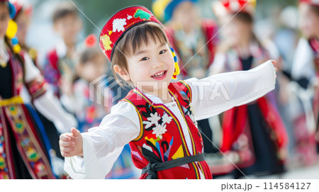 Smiling child in bright traditional attire with arms outstretched during a cultural festival. 114584127