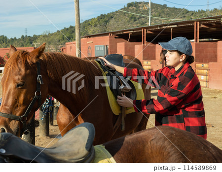 Positive young woman invites to ride horse at horse farm 114589869