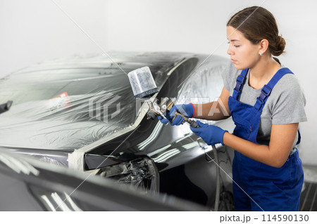 Young female auto painter painting black car using spray gun 114590130