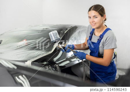 Car painting and automobile repair service. Woman in blue overalls paints car with airbrush pulverizer in paint chamber 114590243