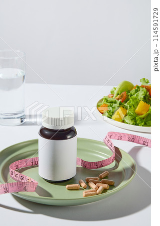 A medicine bottle without label displayed on a round green plate with some yellow pills and a pink measuring tape, a dish of vegetable salad and a glass of water placed in back on white background 114591729