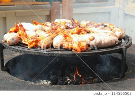 Fried sausages and chicken wings on barbecue grill in the village. Frying meat on open fire. Barbecue party on backyard of country house cabin. Street food festival. Home cooking outdoor. Picnic time Fried sausages and chicken wings on barbecue grill in the village. Frying meat on open fire. Barbecue party on backyard of country house cabin. Street food festival. Home cooking outdoor. Picnic time 114596502