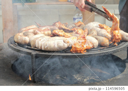 Fried sausages and chicken wings on barbecue grill in the village. Frying meat on open fire. Barbecue party on backyard of country house cabin. Street food festival. Home cooking outdoor. Picnic time Fried sausages and chicken wings on barbecue grill in the village. Frying meat on open fire. Barbecue party on backyard of country house cabin. Street food festival. Home cooking outdoor. Picnic time 114596505
