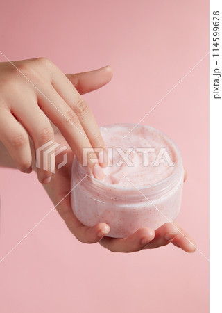 Photo was taken on pink background from front view, a jar of exfoliating scrub may be derived from Himalayan pink salt, a hand is touching into cream texture. Blank space for designing 114599628