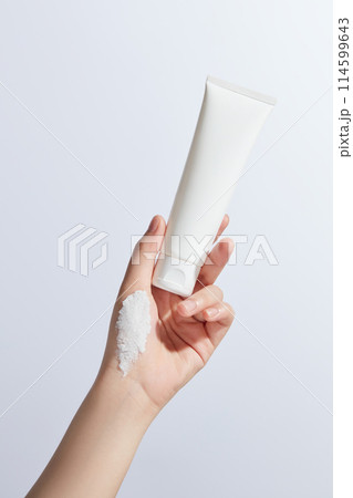 An white blank label cosmetic tube being held by a beautiful, a streak of white cream in hand palm, over white background. Mock up for design with blank space for adding text 114599643
