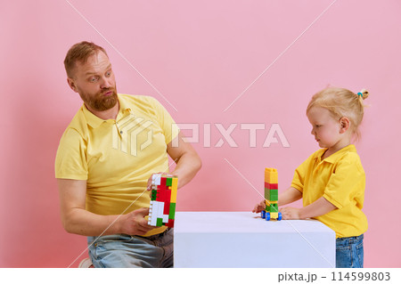 Father and his young son engage in playful building block competition against pink studio background. 114599803