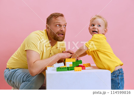 Father guides his little boy, son in building toy with vivid blocks against pink studio background. 114599816