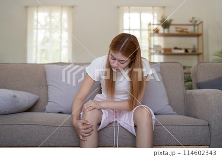 Young Asian woman experiencing knee pain while sitting on a sofa at home. Concept of health issues and discomfort 114602343