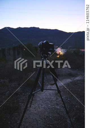 Vertical shot of modern digital camera placed on tripod in nature outdoors, ready for capturing steady pictures or video of a village in mountains at dawn. World photography day concept. Vertical shot of modern digital camera placed on tripod in nature outdoors, ready for capturing steady pictures or video of a village in mountains at dawn. World photography day concept. 114603762