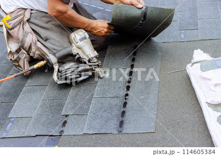 Roofer uses an air pneumatic nail gun to attach new asphalt bitumen shingles to roof Roofer uses an air pneumatic nail gun to attach new asphalt bitumen shingles to roof 114605384