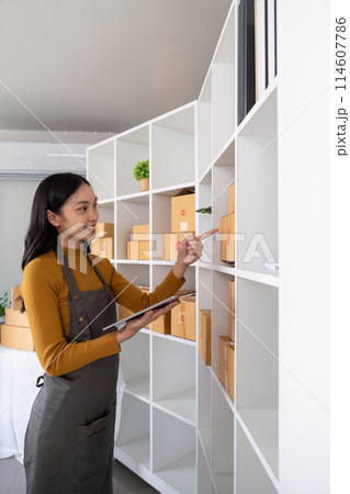 Ecommerce business owner managing inventory and checking stock in home office. Small business entrepreneur organizing packages for shipment and using a tablet for inventory tracking Ecommerce business owner managing inventory and checking stock in home office. Small business entrepreneur organizing packages for shipment and using a tablet for inventory tracking 114607786