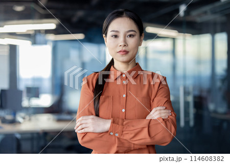 A serious thinking Asian businesswoman in a modern office. She conveys professionalism and confidence with arms crossed, reflecting the work environment. A serious thinking Asian businesswoman in a modern office. She conveys professionalism and confidence with arms crossed, reflecting the work environment. 114608382