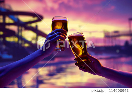 Close-up of hands toasting with beer glasses at sunset, with a amusement park and water slides blurred in the background. 114608941