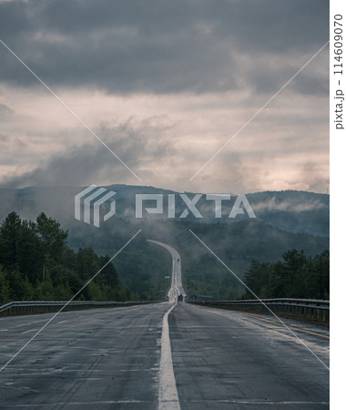 Straight empty highway stretching toward distant forest on a cloudy day Straight empty highway stretching toward distant forest on a cloudy day 114609070