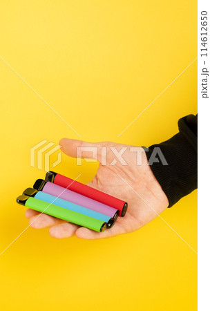 Disposable electronic cigarettes of different flavors in hand on a yellow background. The concept of modern smoking, vaping and nicotine. Disposable electronic cigarettes of different flavors in hand on a yellow background. The concept of modern smoking, vaping and nicotine. 114612650
