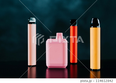 A set of colorful disposable electronic cigarettes of different shapes on a black background with smoke. Concept of modern smoking, vaping and nicotine. 114612842