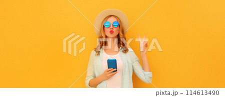 Stylish woman posing blowing a kiss with smartphone wearing summer hat posing on yellow background 114613490