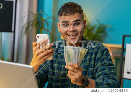 Smiling happy man counting money dollar cash use smartphone income saves lottery win at home Smiling happy man counting money dollar cash use smartphone income saves lottery win at home 114614389