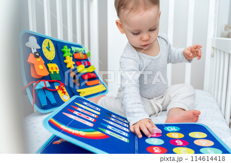 Little toddler girl playing with busy book while sitting in crib. Concept of quiet books and modern educational toys for kids Little toddler girl playing with busy book while sitting in crib. Concept of quiet books and modern educational toys for kids 114614418