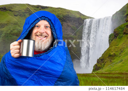 bearded man in a sleeping bag with a cup of coffee against the backdrop of nature in the mountains 114617484