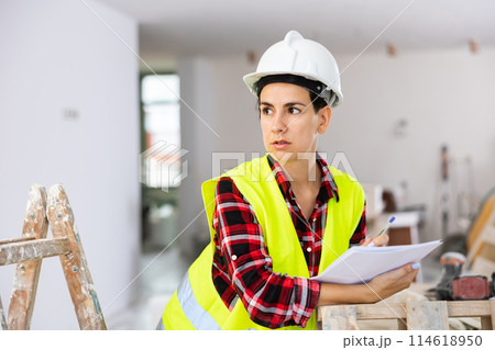 Portrait of excited young woman builder next to stepladder with documents in her hands checks quality of completed repairs 114618950