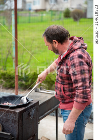 Mature russian man frying meat in a cauldron on the stove outdoors, he stirs meat with a spatula 114619523