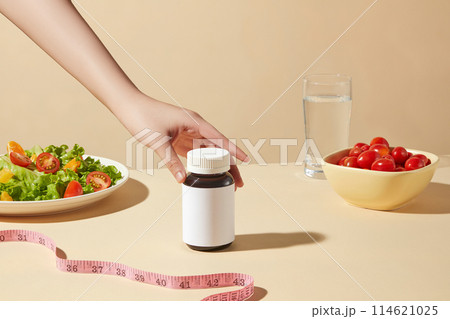 Template for weight losing product advertising, a medicine bottle without label placed in the middle of table is touched by a hand, surrounded by healthy foods for dietary. Front view, copy space Template for weight losing product advertising, a medicine bottle without label placed in the middle of table is touched by a hand, surrounded by healthy foods for dietary. Front view, copy space 114621025