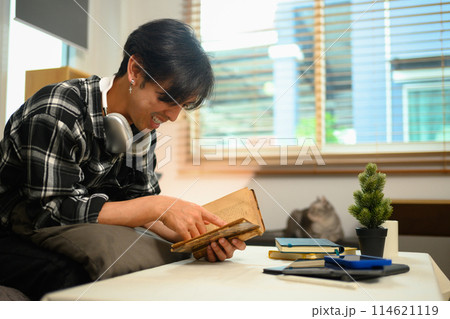Carefree young Asian man sitting on couch in living room and reading book Carefree young Asian man sitting on couch in living room and reading book 114621119