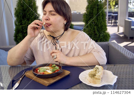 medium sized woman in peach fuzz dress eating Shakshouka in modern restaurant 114627304