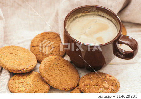 A cup of aromatic coffee cappuccino with Oatmeal cookies. Morning rays and latte for breakfast in a beautiful brown cup. Wooden background. A cup of aromatic coffee cappuccino with Oatmeal cookies. Morning rays and latte for breakfast in a beautiful brown cup. Wooden background. 114629235
