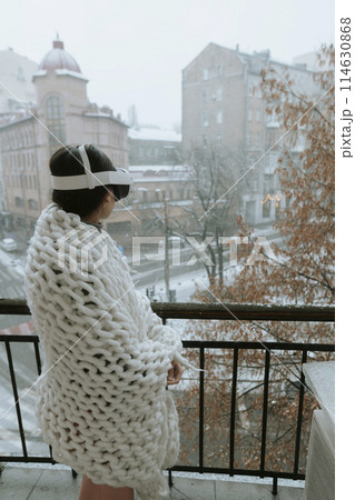 Amidst snowy landscapes, a young lady enjoys VR on the balcony with a headset. 114630868