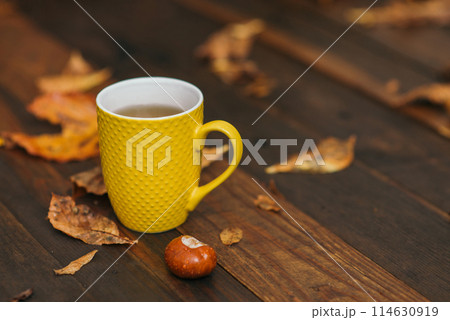 Yellow cup of tea and chestnut at wooden background with a lot of red and yellow fallen leaves. Relax scene. Selective focus and copy space. 114630919