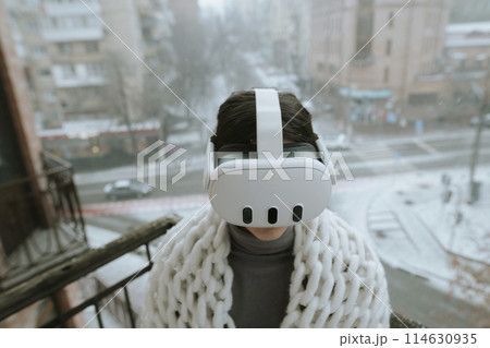 On the balcony in winter, a girl embraces virtual reality adventures with a headset. On the balcony in winter, a girl embraces virtual reality adventures with a headset. 114630935