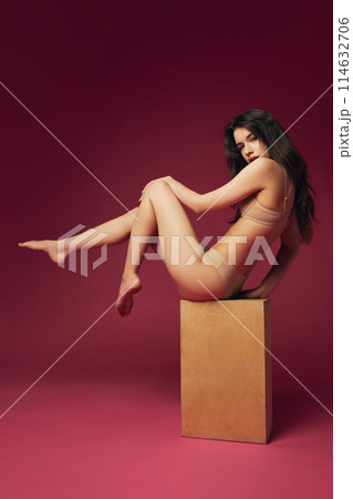 Beautiful woman with brunet hair, slim fit body posing on cube in beige underwear against magenta studio background Beautiful woman with brunet hair, slim fit body posing on cube in beige underwear against magenta studio background 114632706