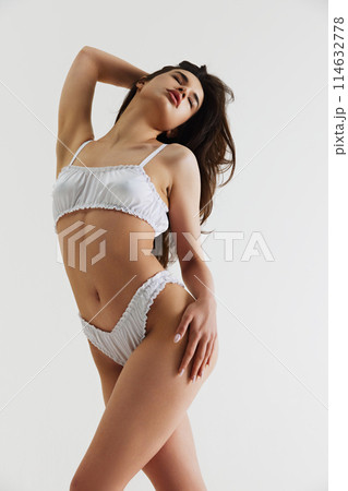 Sensuality and femininity vibe. Beautiful young woman in white elegant lingerie, with slim body posing isolated on grey studio background Sensuality and femininity vibe. Beautiful young woman in white elegant lingerie, with slim body posing isolated on grey studio background 114632778