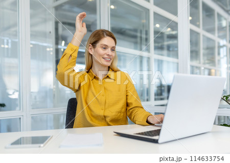 Smiling businesswoman celebrating success during an online meeting at the office desk. She is raising her hand with joy while working on her laptop. 114633754