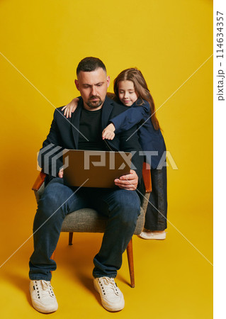Father working online and his little daughter dressed formal attire pointing to screen against sunny-yellow background. 114634357